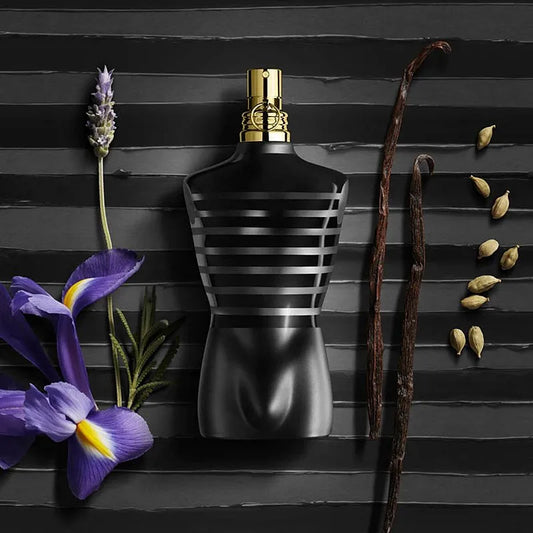 Black striped Jean Paul Le Male perfume bottle with gold cap on a black background with flowers and vanilla beans.